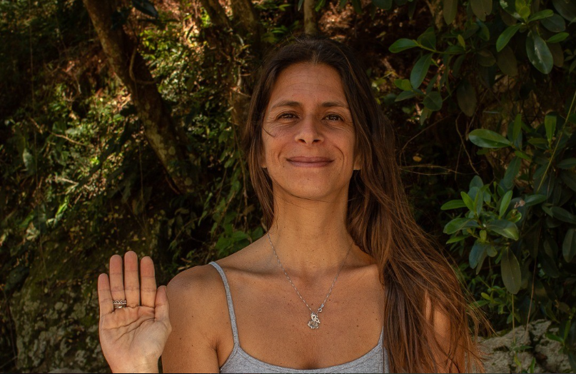 Ana Lima sorrindo em meio à mata, com a mão erguida em gesto de saudação